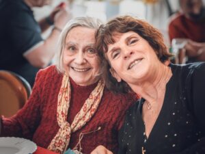 Deux femmes souriantes, une âgée et une plus jeune, assises côte à côte lors d’un repas convivial.