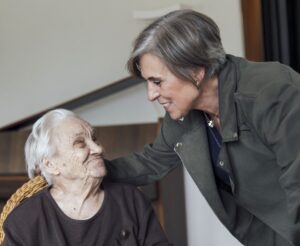 Une femme penchée vers une dame très âgée assise dans un fauteuil en osier, lui pose la main sur l'épaule avec tendresse, leurs regards se croisant dans un échange complice.