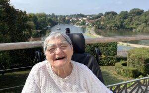 Une femme âgée souriante, assise dans son fauteuil roulant, profite d'un beau panorama sur une rivière bordée de verdure et de maisons.