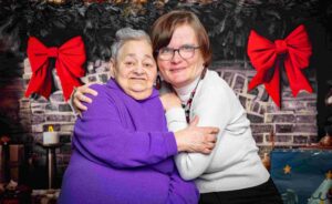 Deux femmes souriantes, l’une âgée et l’autre plus jeune, posent enlacées devant une cheminée décorée de Noël avec des rubans rouges.
