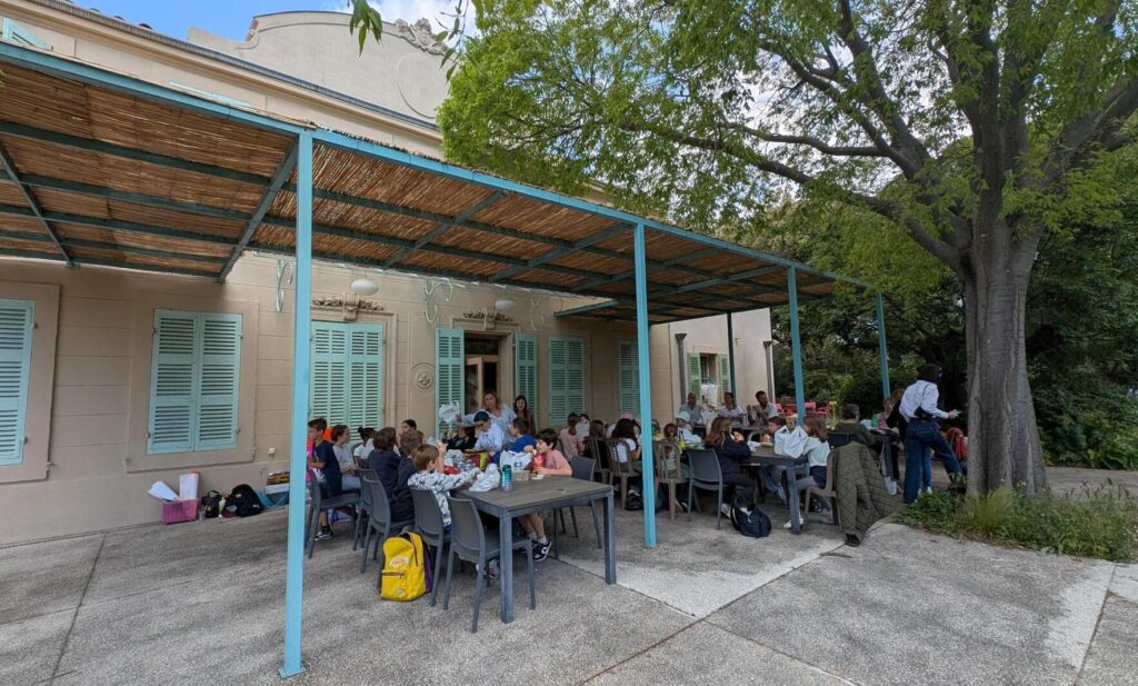 Photo de la journée intergénérationnelle 2025 à Marseille avec la classe de CM1 au complet et les personnes âgées accompagnées sous un préau en train de déjeuner.
