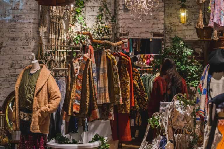 Photo d’une friperie vintage avec des vêtements colorés sur portants, un mannequin habillé et une cliente en train de regarder les articles.