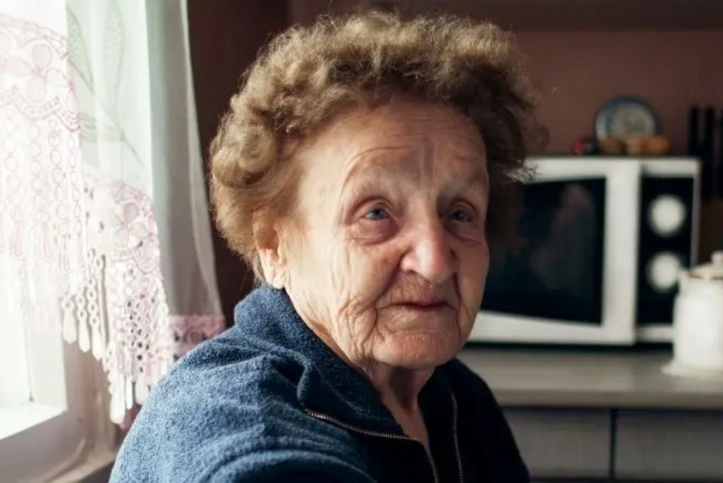 Portrait d’une femme âgée, assise chez elle, regardant vers l’objectif, le visage marqué par le temps.