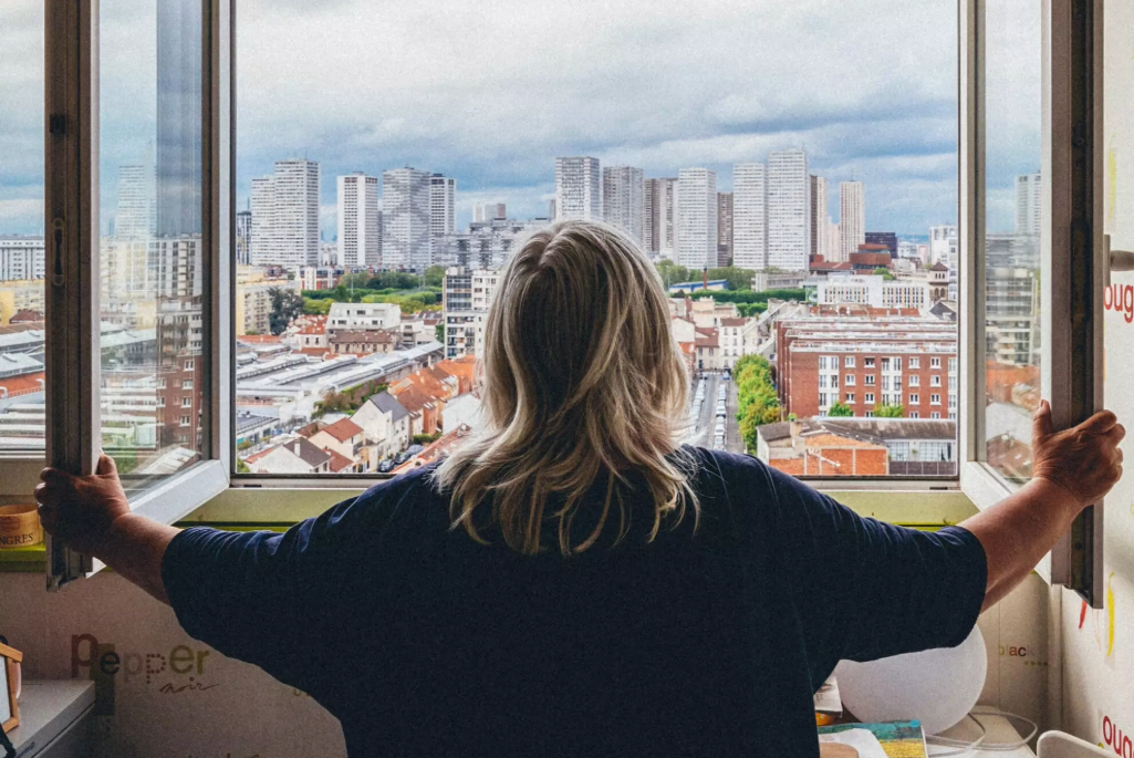 Une femme âgée, de dos, ouvre ses fenêtres sur une vue urbaine avec des immeubles en arrière-plan.