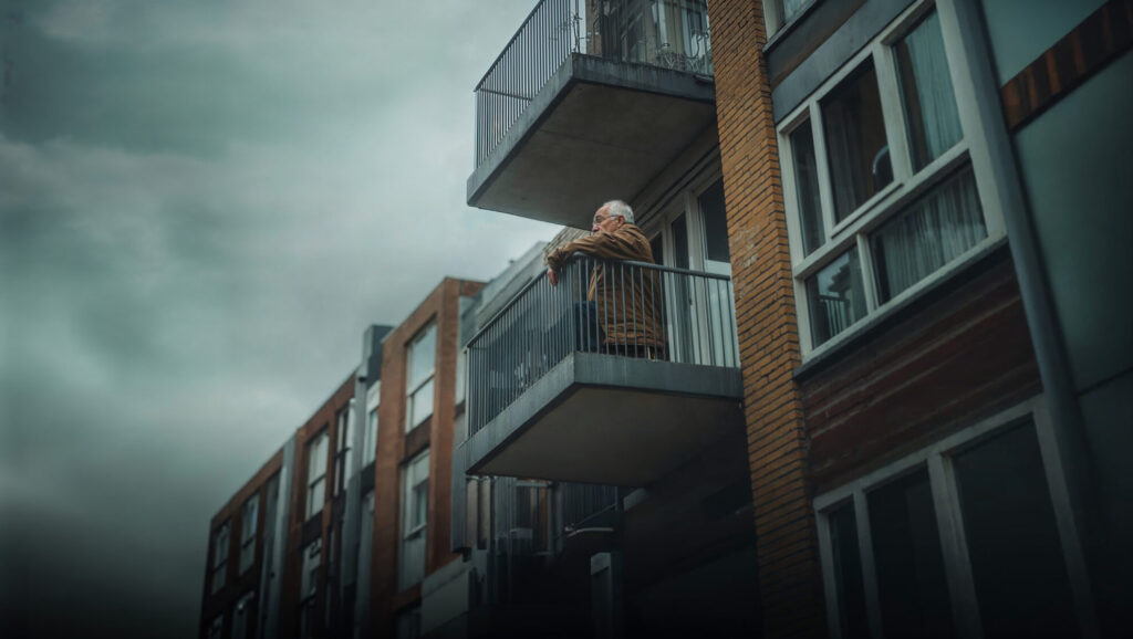 Un homme âgé seul sur le balcon de son appartement en immeuble, regardant dehors.