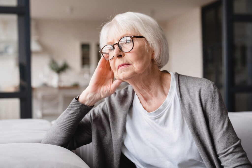 Femme âgée au regard pensif, assise chez elle, la main posée contre sa tempe, exprimant une forme de solitude ou de mélancolie.
