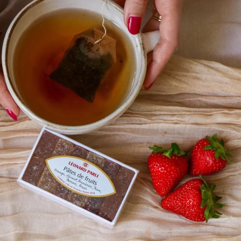 Tasse de thé avec sachet infusé, boîte de pâtes de fruits Léonard Parli et trois fraises fraîches posées sur un tissu beige.
