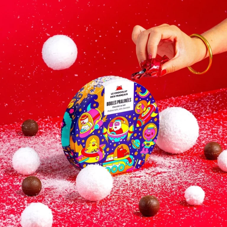 Coffret coloré de boules pralinées « Le Chocolat des Français » posé sur un fond rouge avec des boules de neige et des chocolats dispersés, une main ouvre une papillote brillante.