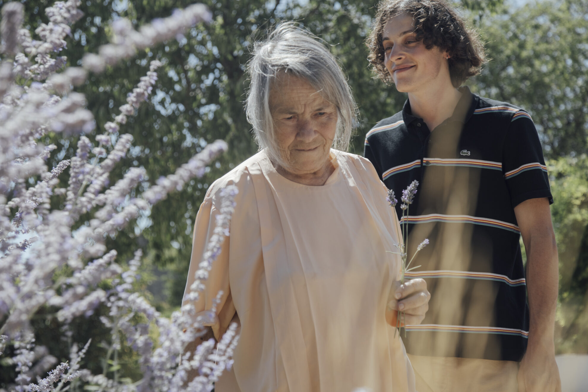 Femme âgée tenant un brin de lavande dans un jardin ensoleillé, avec un jeune homme souriant derrière elle.