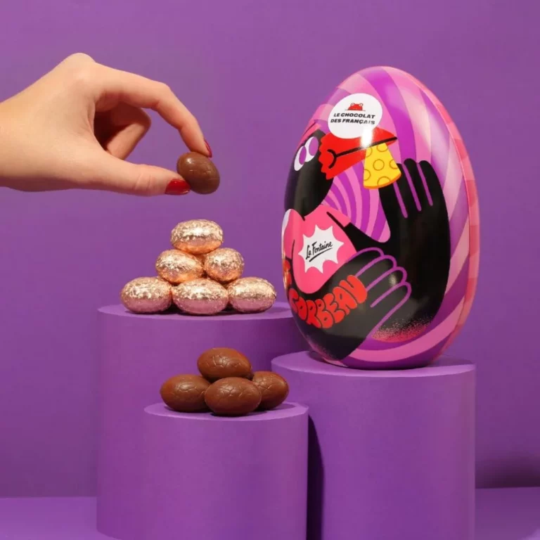 Main prenant un œuf en chocolat devant une boîte métallique en forme d’œuf décorée posée sur des socles violets.