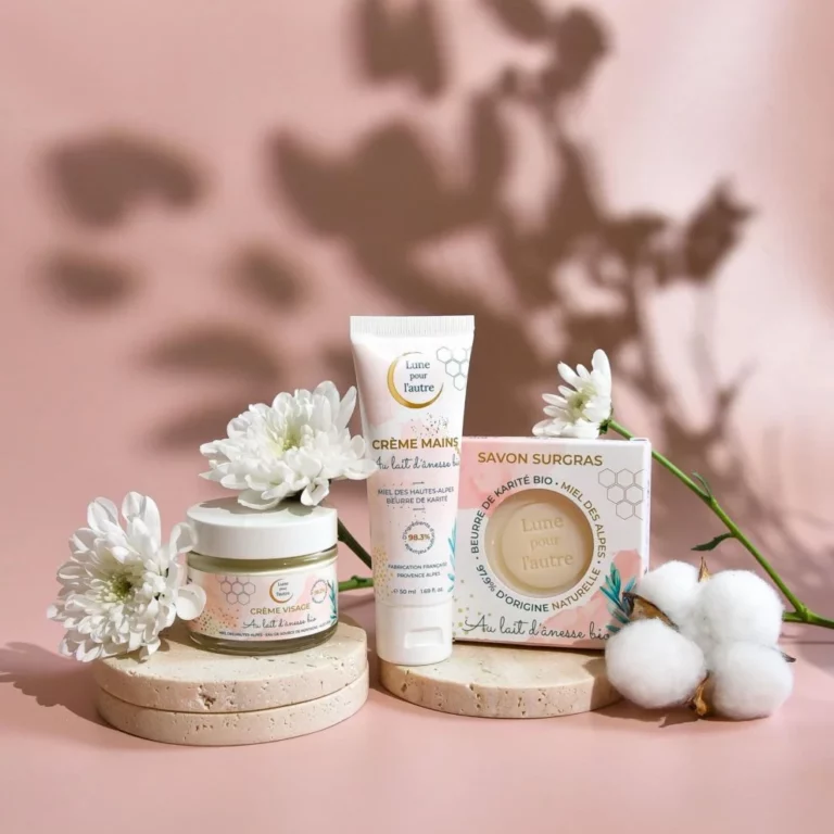 Coffret de soins au lait d’ânesse comprenant une crème visage, une crème mains et un savon, disposés avec des fleurs sur fond rose.