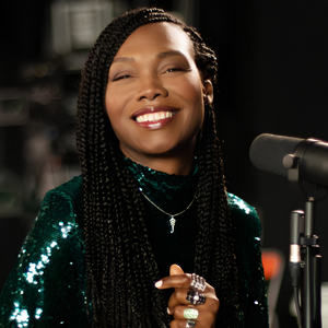 Artiste souriante portant une tenue à paillettes verte devant un microphone dans un studio sombre.