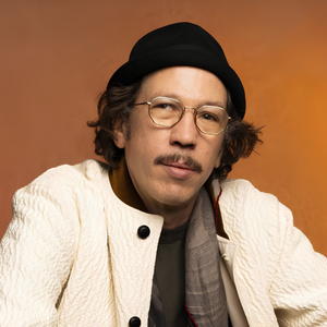 Portrait d’un homme portant un chapeau noir, des lunettes et une veste blanche texturée devant un fond orange.