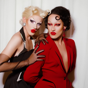 Deux personnes maquillées et vêtues de tenues élégantes, l’une en costume rouge et l’autre en noir et blanc, posant devant un fond clair.