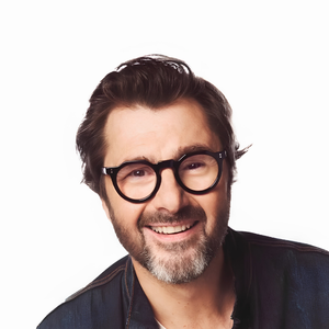 Portrait d’un homme souriant portant des lunettes rondes et une veste sombre devant un fond blanc.