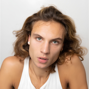 Portrait rapproché d’un artiste aux cheveux longs portant un débardeur blanc devant un fond clair.