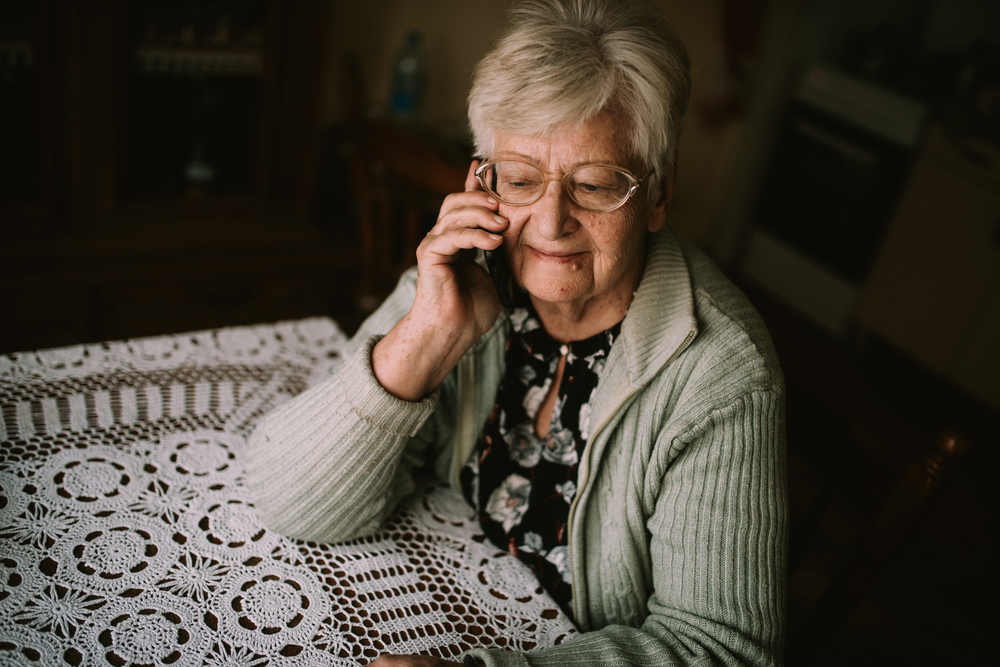 Personne âgée souriante répondant au téléphone dans un intérieur frais, volets mi-clos, pendant un épisode de canicule