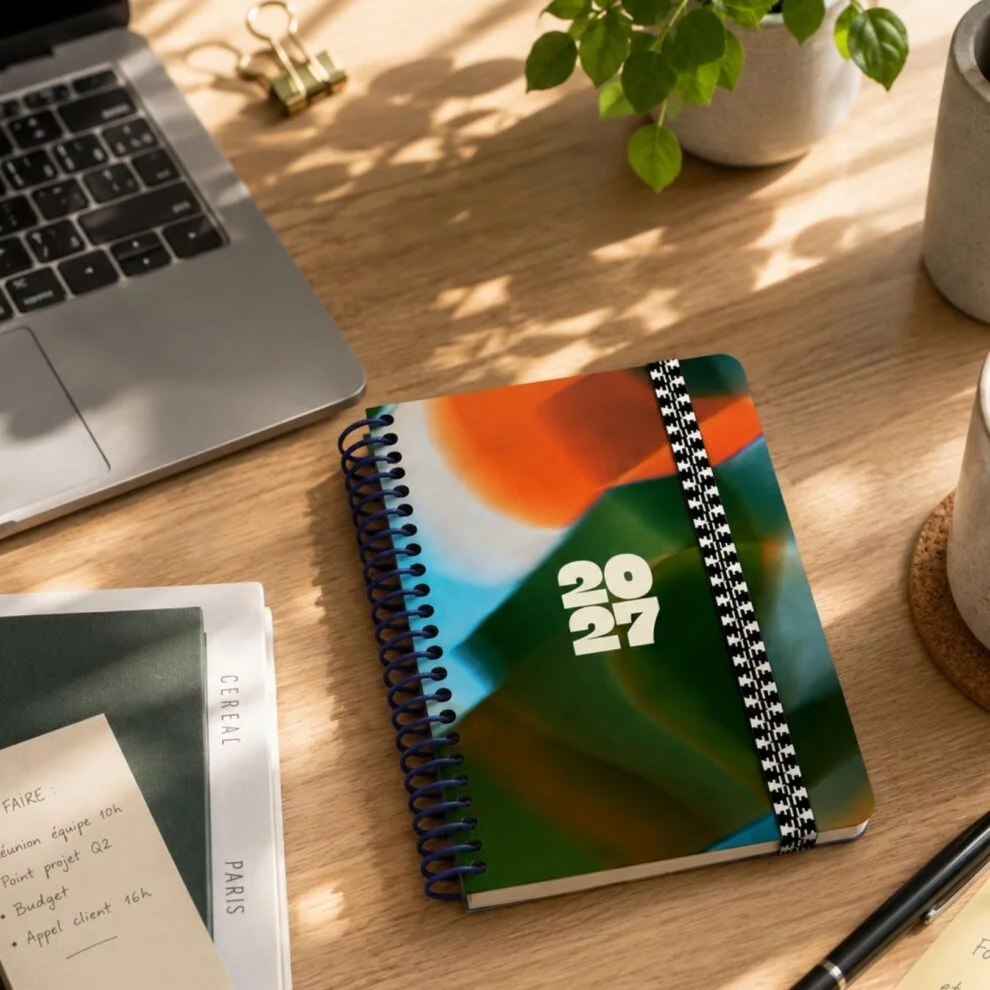 Agenda spiralé 2027 à couverture abstraite orange, vert et bleu, posé sur un bureau en bois à côté d'un ordinateur portable et d'une plante verte.