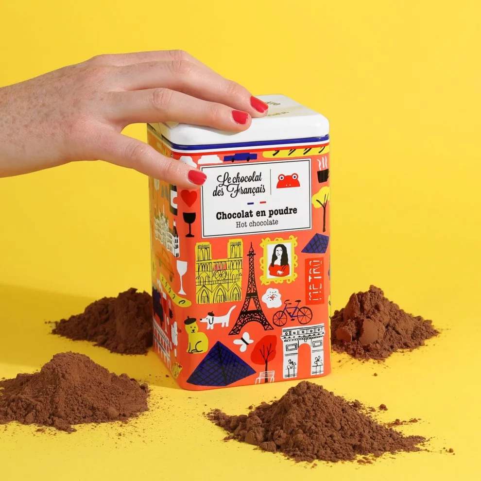 Une main ouvre une boîte métallique illustrée de monuments parisiens, « Le chocolat des Français », entourée de tas de chocolat en poudre, sur fond jaune.